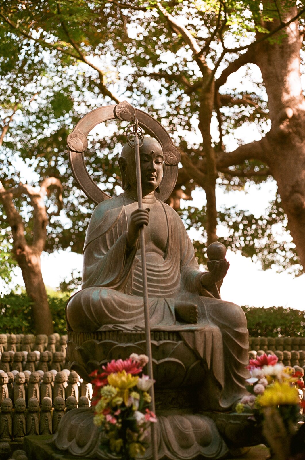 Statue assise tenant un bâton, sous des arbres en contre-jour